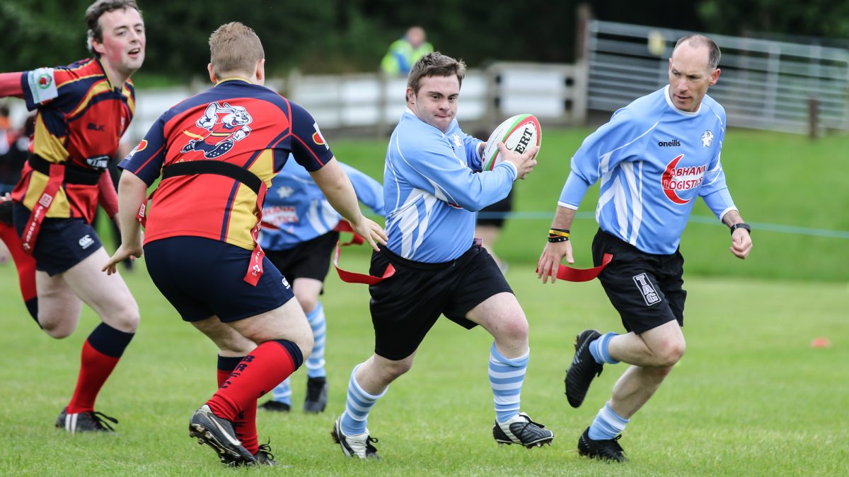 boy in blue runs with Gilbert branded rugby ball from competitors in red in game of tag rugby