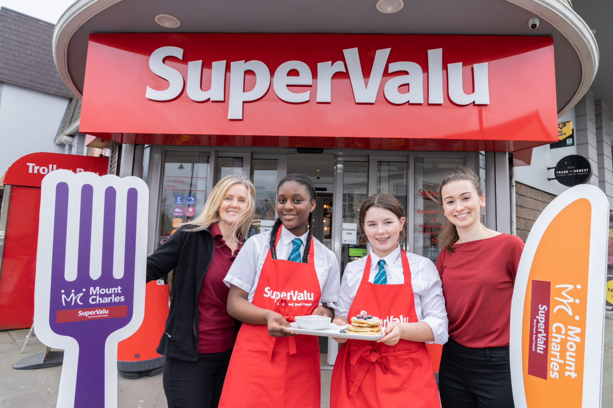 Students pose with tray of pancakes and novelty human size cutlery in front of SuperValu Comber as part of cooking competition