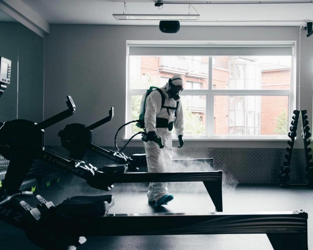 Mount Charles cleaning team member wearing a white chemical safe suit, with black mask spraying sanitising chemicals on sports equipment