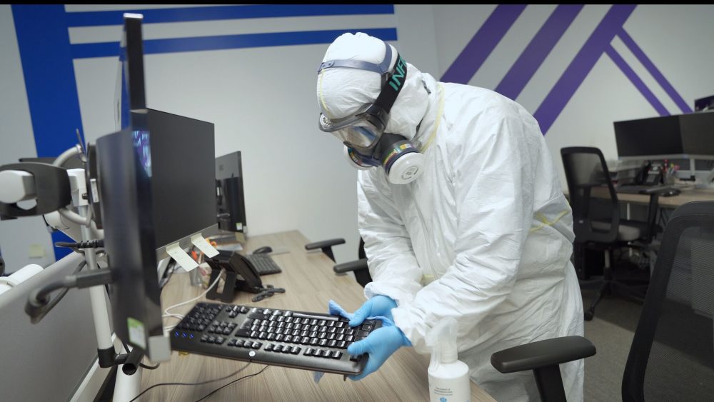 Mount Charles team member wearing a white hazardous safe suit with black mask cleaning computer equipment in an office with sanitising chemicals