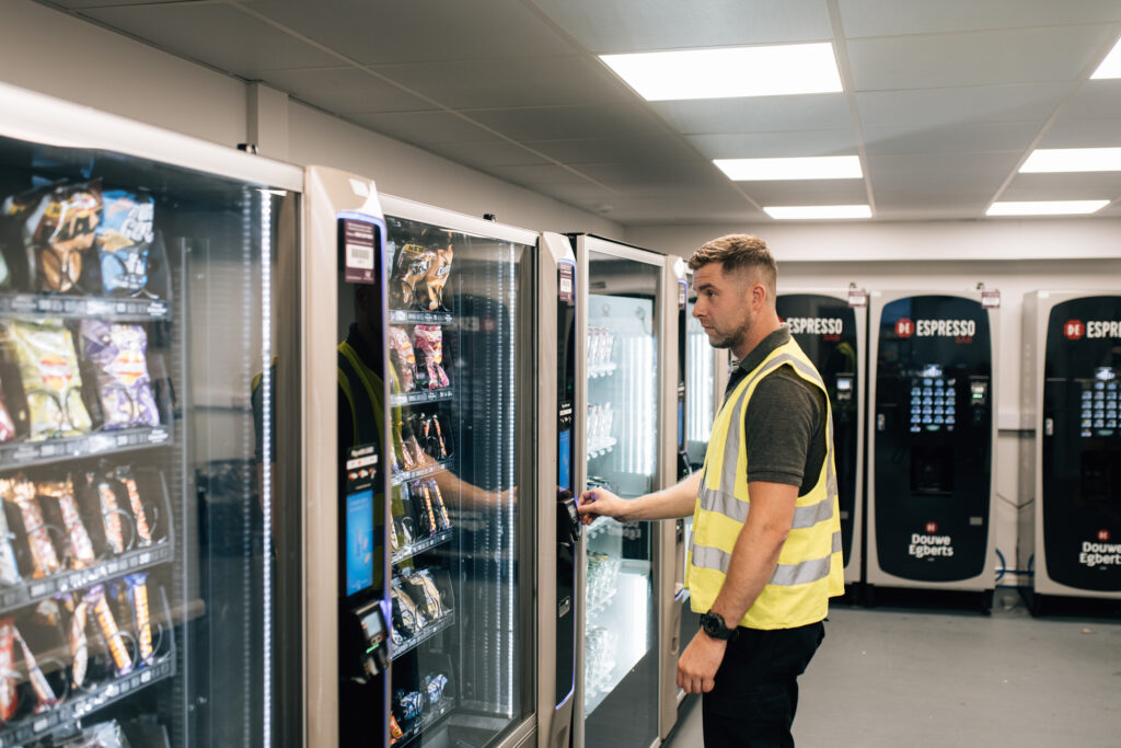 A man in high vis purchases items from a vending machine, in an area where there are several machines selling snacks and also coffee