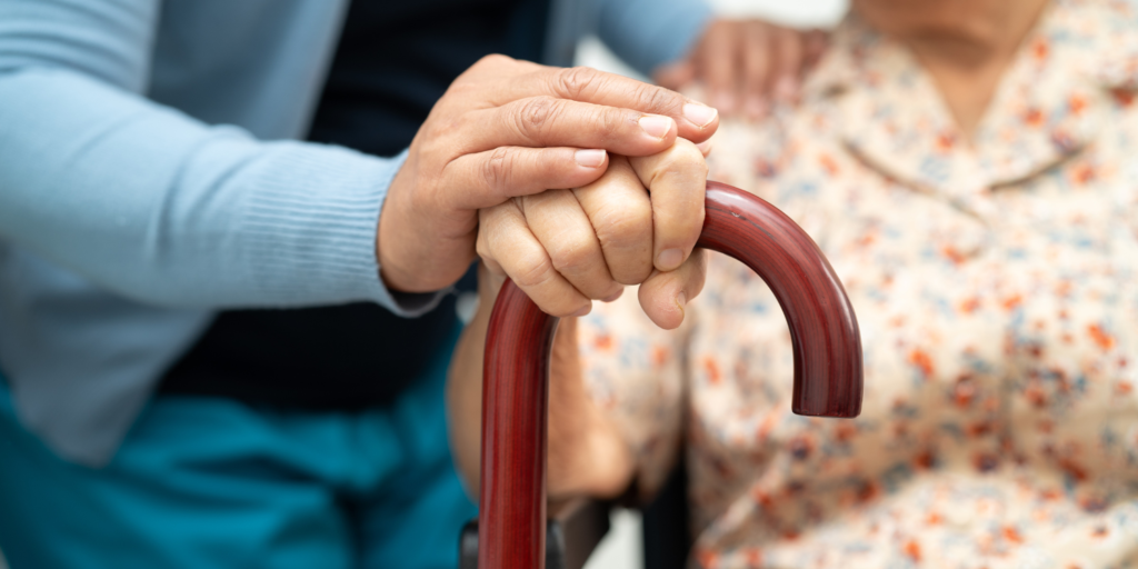 A carer and elderly patient in the background with their hands leaning on the patient's walking stick in the foreground