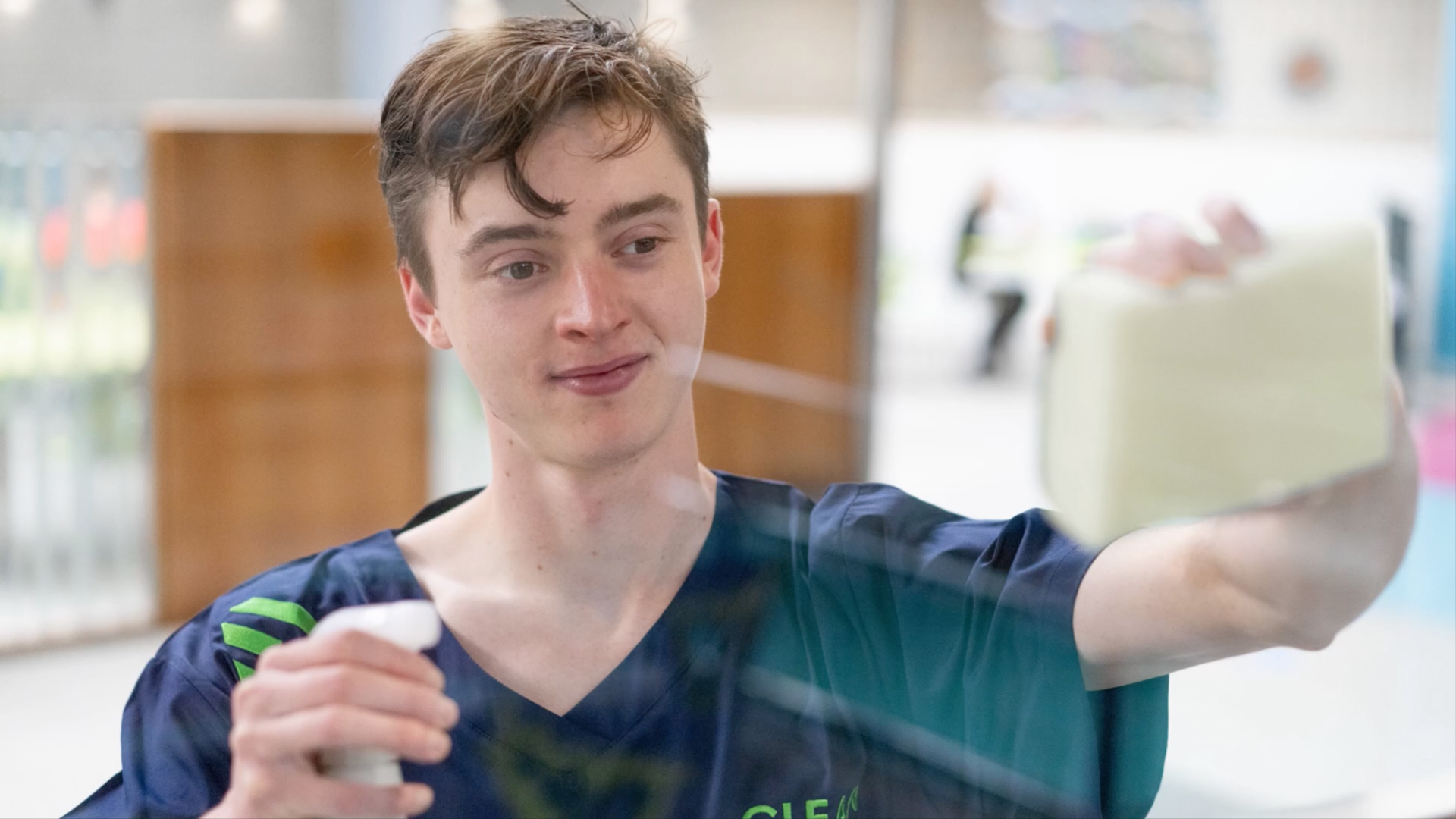 A Cleaning Operative uses sponge and spray to clean windows at MC site, wearing MC Clean uniform